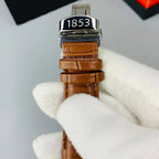 Watch with brown strap and '1853' branding held by a gloved hand on a gray background.
